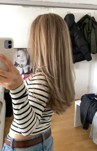 Eine Person von hinten mit langem, glattem, hellbraunem Haar und natürlichen hellen Highlights im Spiegelbild.