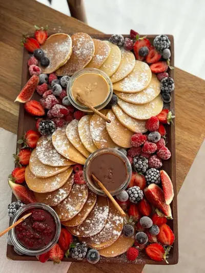 Ein Holzbrett mit kleinen Pfannkuchen, frischen Beeren, Feigen und drei verschiedenen Dips in kleinen Glasschalen.