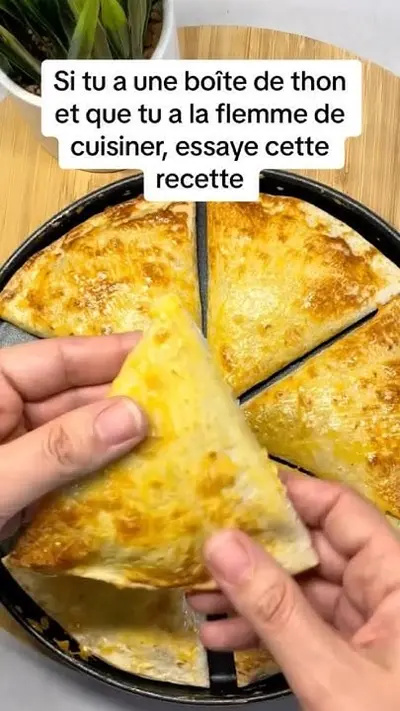 Une main tient un triangle de tortilla doré garni de thon et de fromage fondu au-dessus d'une poêle.