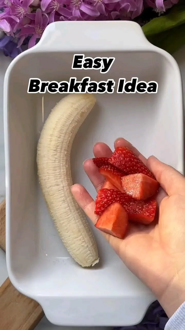 Eine Hand gibt geschnittene Erdbeeren zu einer geschälten Banane in eine weiße Auflaufform.