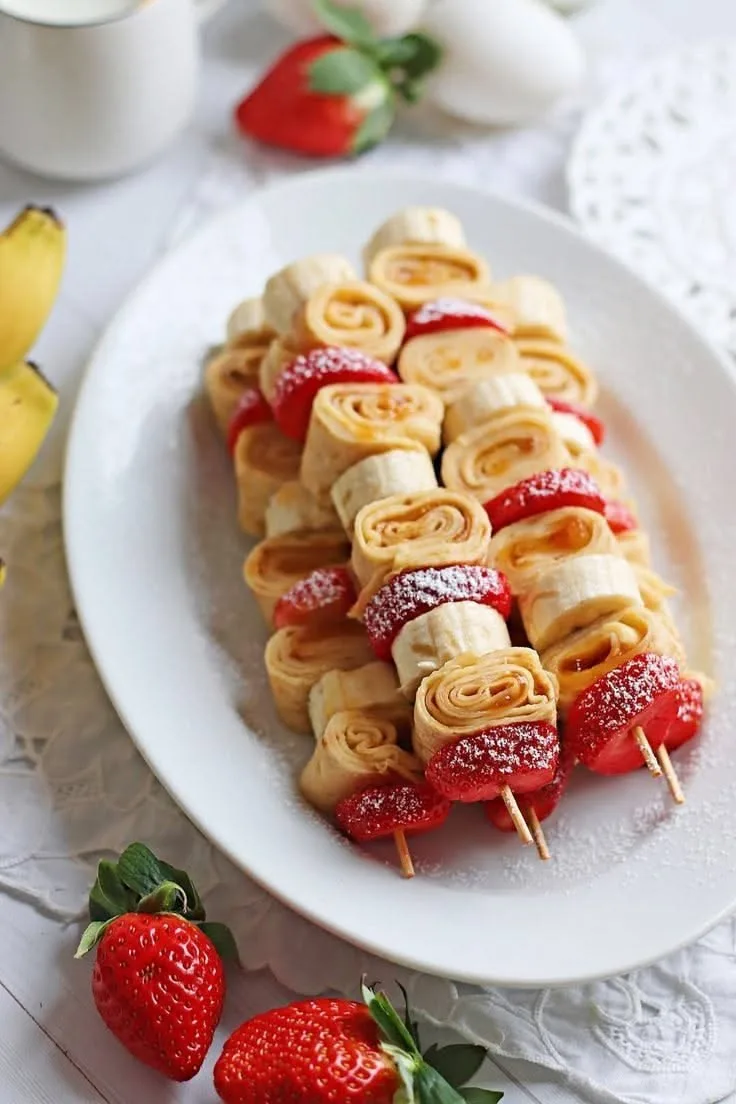 Mehrere Holzspieße mit gerollten Crepe-Stücken, Bananenscheiben und Erdbeeren auf einer weißen Servierplatte.