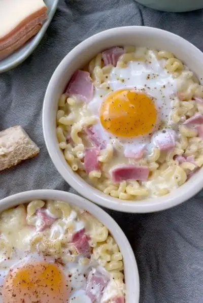 Deux bols de macaronis gratinés au jambon surmontés d'un œuf au plat, servis sur une nappe grise.