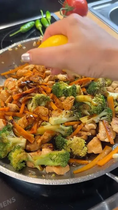 Une main presse un citron sur une poêlée de poulet, de brocolis et de carottes dans une poêle.