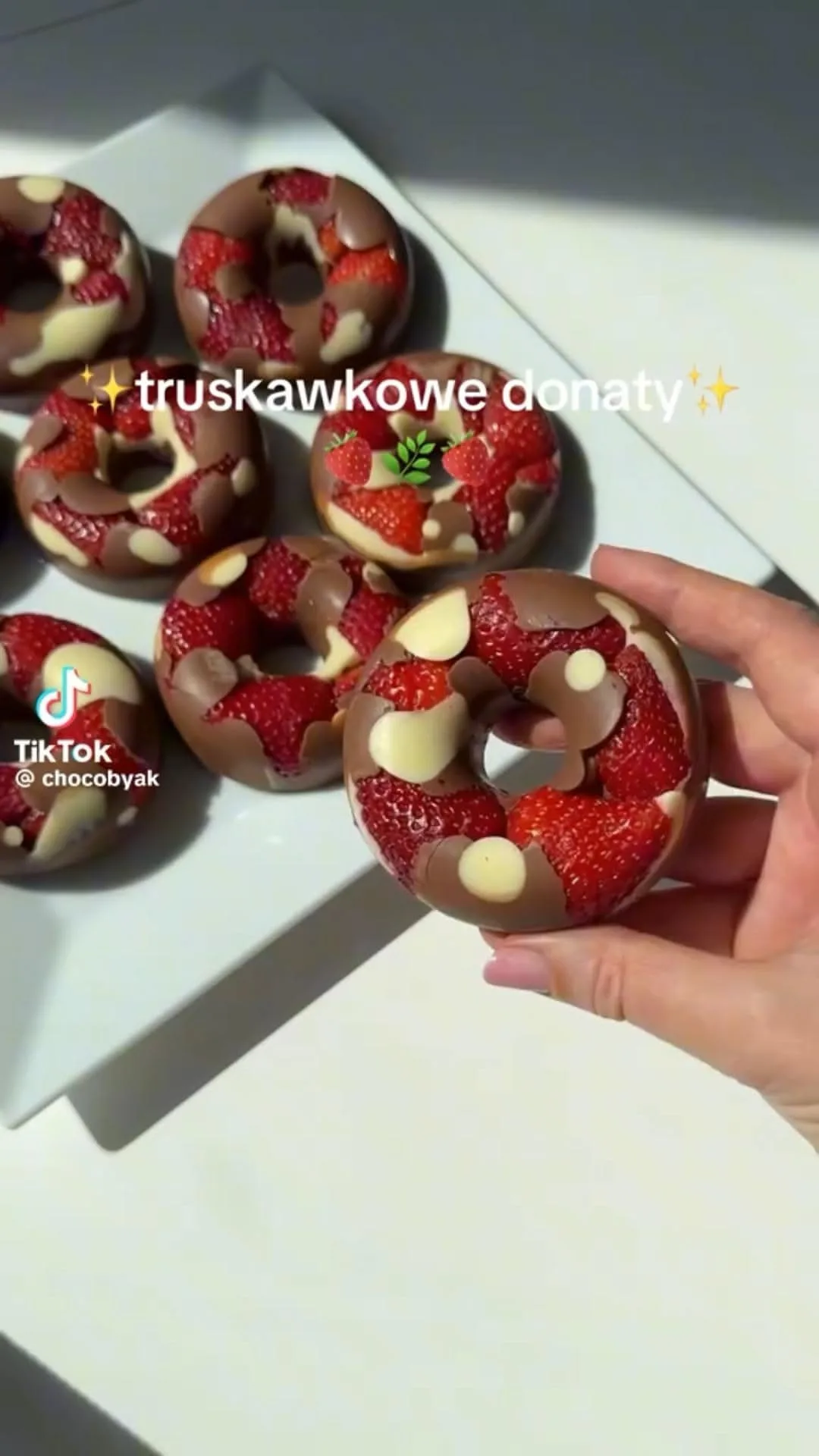 Eine Hand hält einen mit Erdbeeren und Schokolade verzierten Donut über einem Teller mit weiteren Donuts.