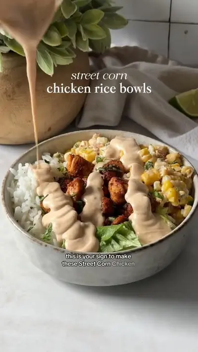 Een kom gevuld met rijst, gegrilde kip, maïs en sla waarover een romige dressing wordt gegoten.