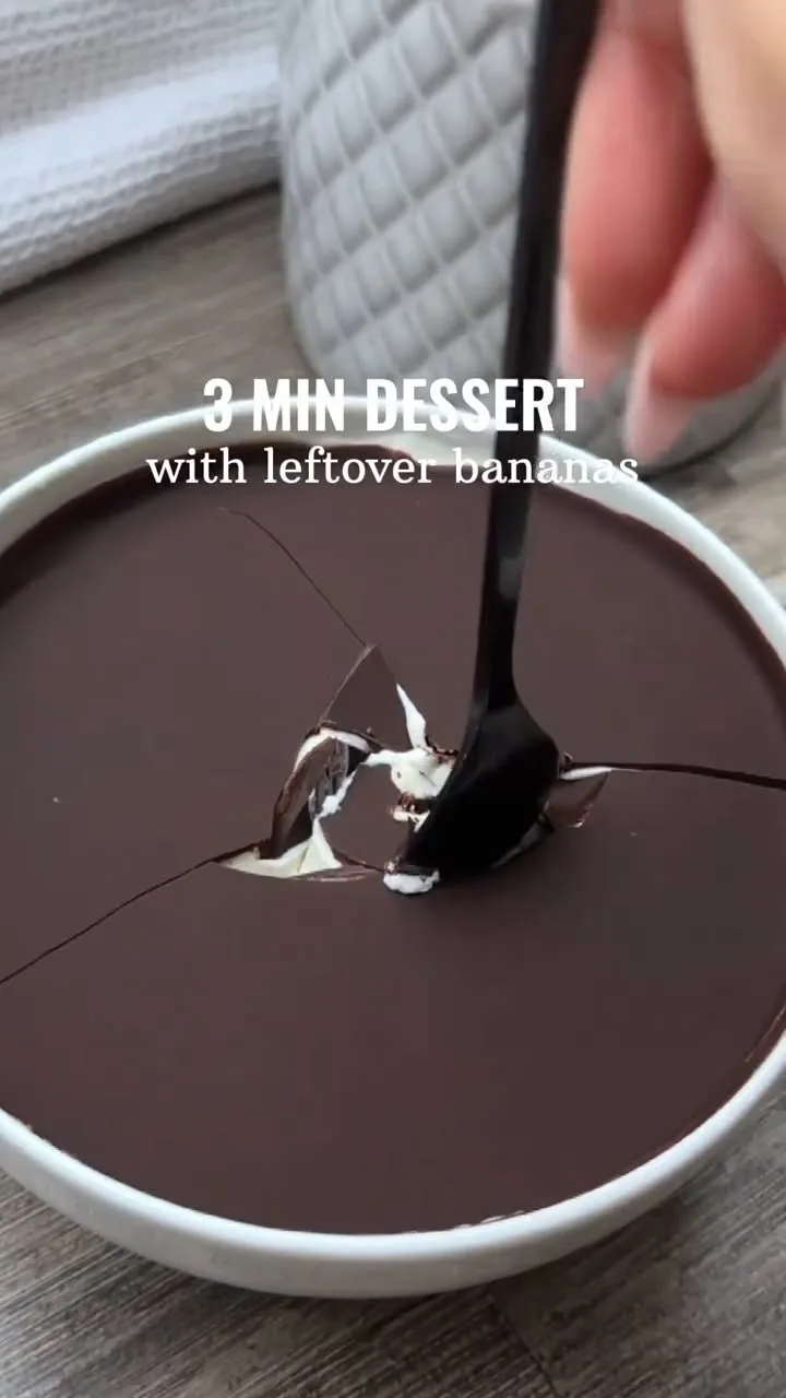 Une cuillère noire brise une couche de chocolat noir craquant dans un bol, dévoilant une crème de banane onctueuse.