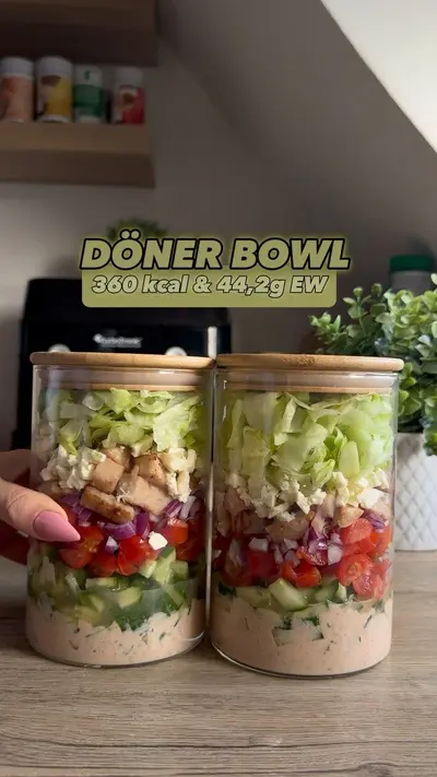 Zwei Glasbehälter mit geschichteten Döner-Bowl-Zutaten von der Soße bis zum Eisbergsalat stehen auf einem Holztisch.
