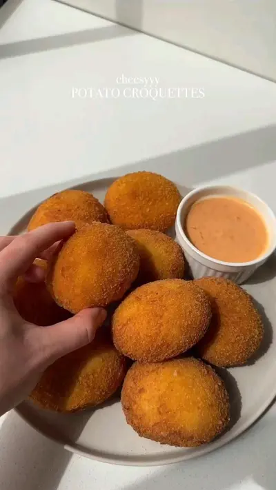 Une main saisit une croquette de pomme de terre dorée au-dessus d'une assiette remplie de bouchées frites et d'une sauce.
