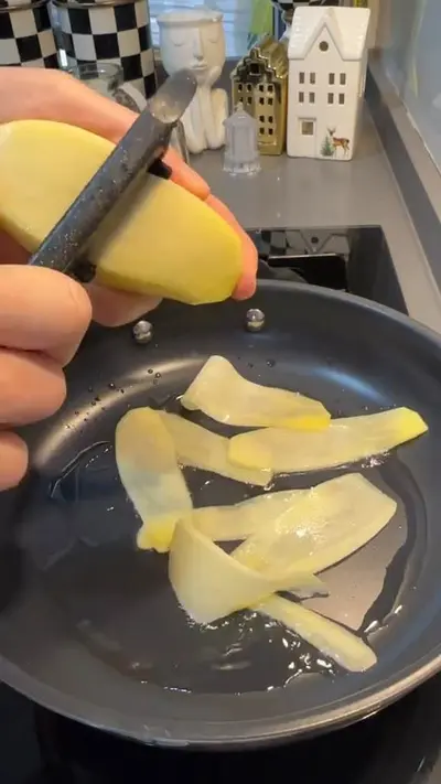 Eine Hand schält eine Kartoffel mit einem Sparschäler direkt in eine Pfanne mit heißem Öl.