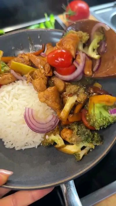 Une poêle contenant du poulet sauté aux brocolis, tomates cerises et oignons rouges, accompagnée de riz blanc.