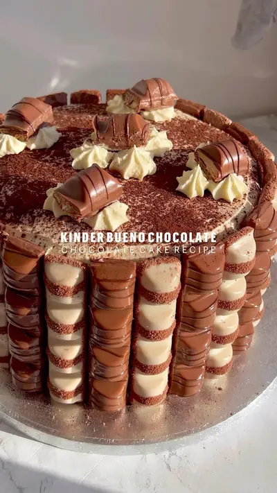 Un gâteau rond recouvert de crème, entouré de barres Kinder Bueno et décoré de cacao en poudre et de morceaux de chocolat.