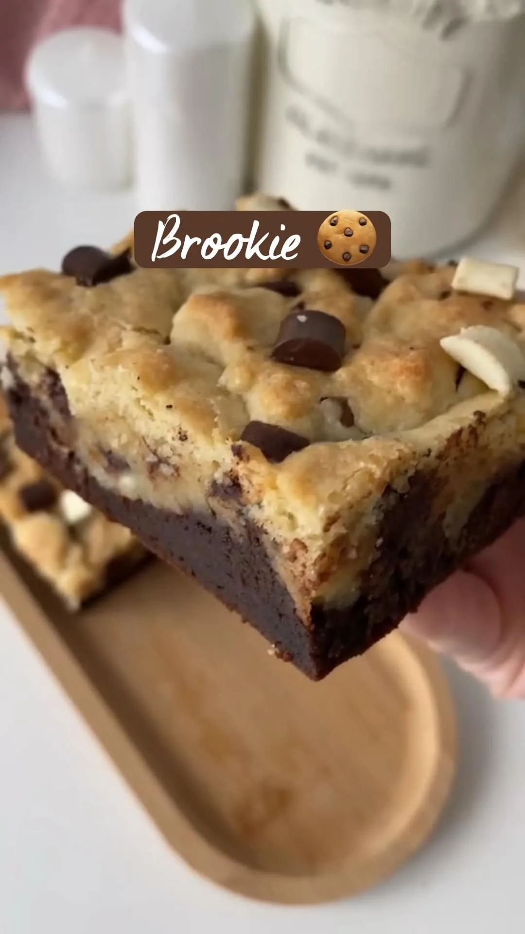 Eine Hand hält ein quadratisches Stück Brookie mit dunkler Brownie-Basis und heller Cookie-Schicht mit Schokostückchen.