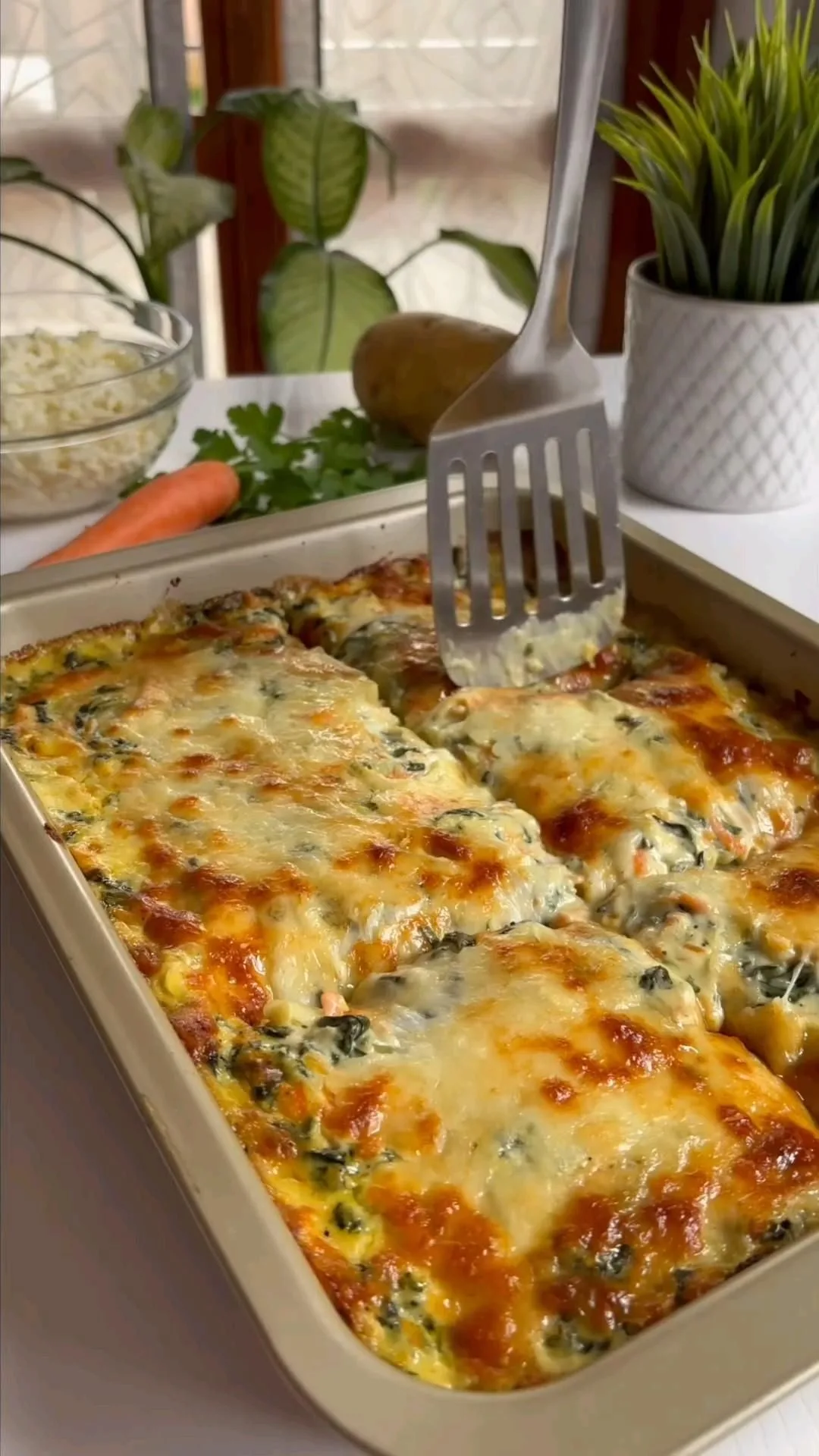 Une spatule soulève une portion de gratin doré aux pommes de terre et épinards dans un plat rectangulaire.
