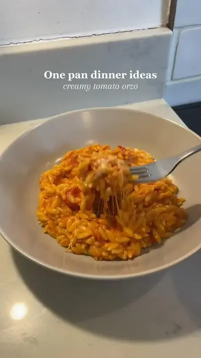 Een close-up van een kom met romige orzo in tomatensaus, waarbij een vork draden gesmolten kaas omhoog tilt.