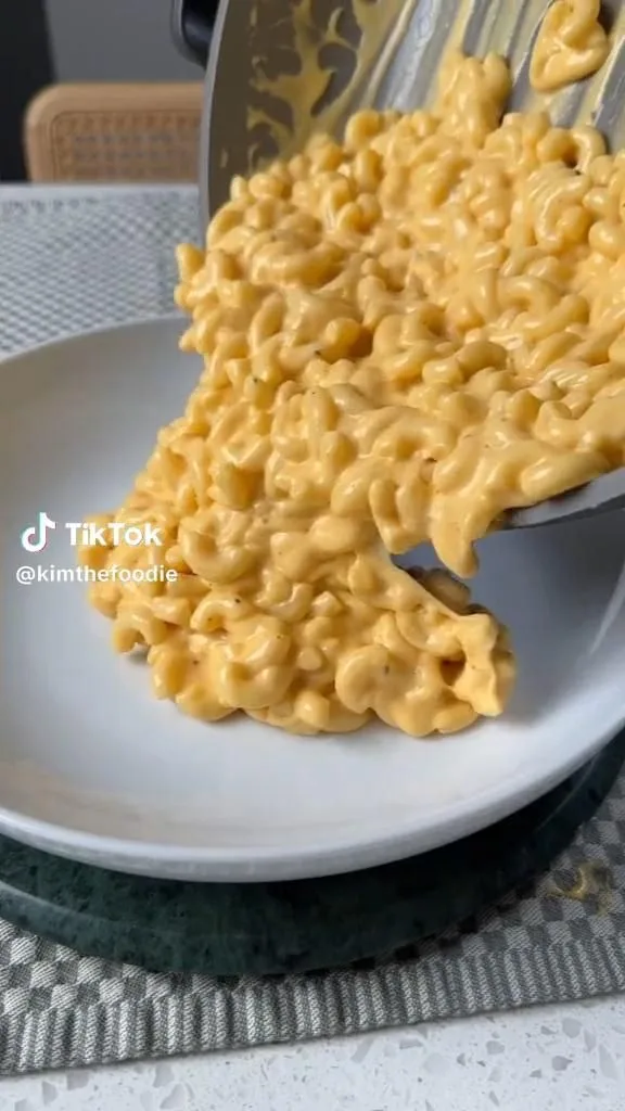 Une poêle verse des macaronis au fromage onctueux et bien jaunes dans une assiette blanche sur un dessous de plat.