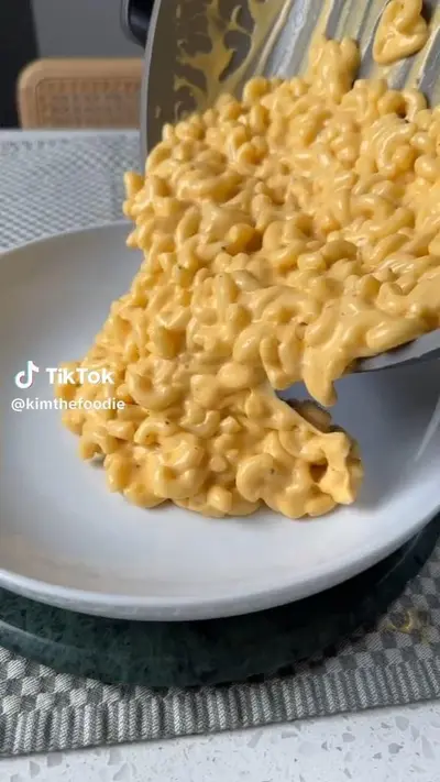Une poêle verse des macaronis au fromage onctueux et bien jaunes dans une assiette blanche sur un dessous de plat.