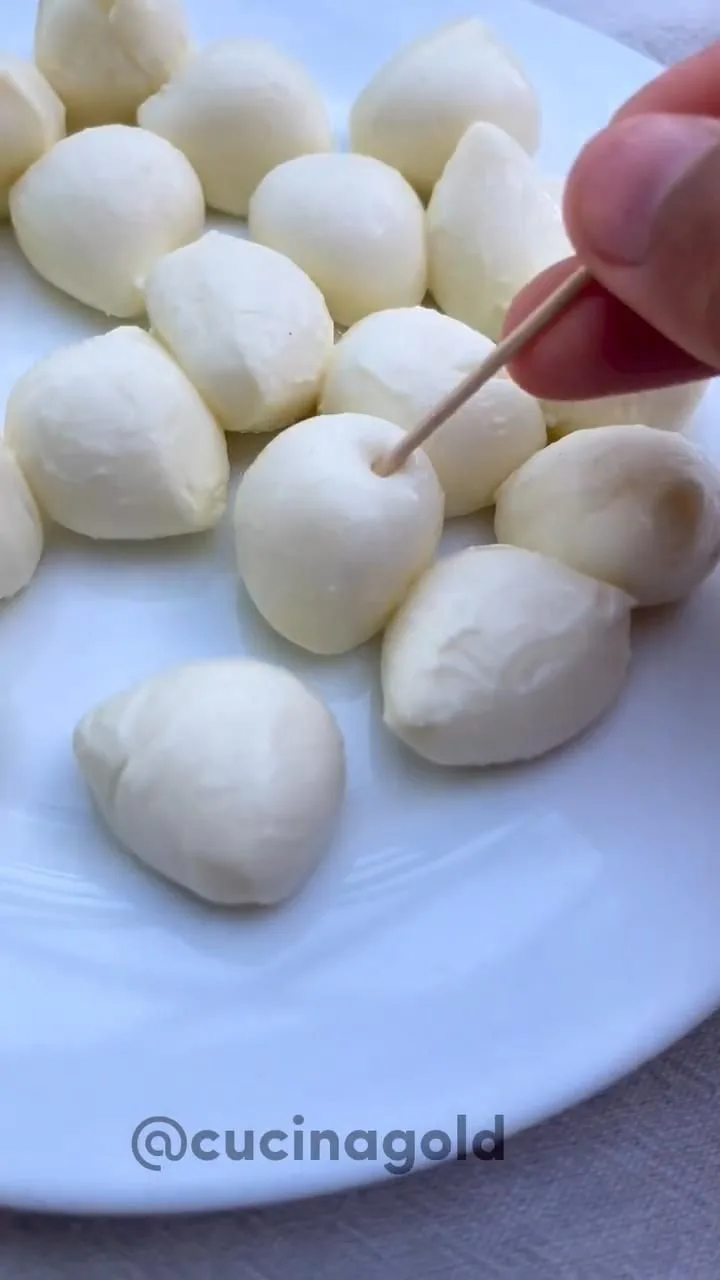 Eine Hand spießt mit einem Zahnstocher eine kleine Mozzarella-Kugel auf einem weißen Teller auf.