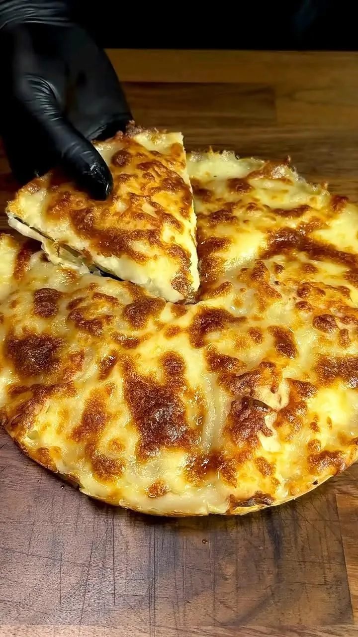 Une main gantée soulève une part de pizza ronde généreusement garnie de fromage fondu sur une planche en bois.