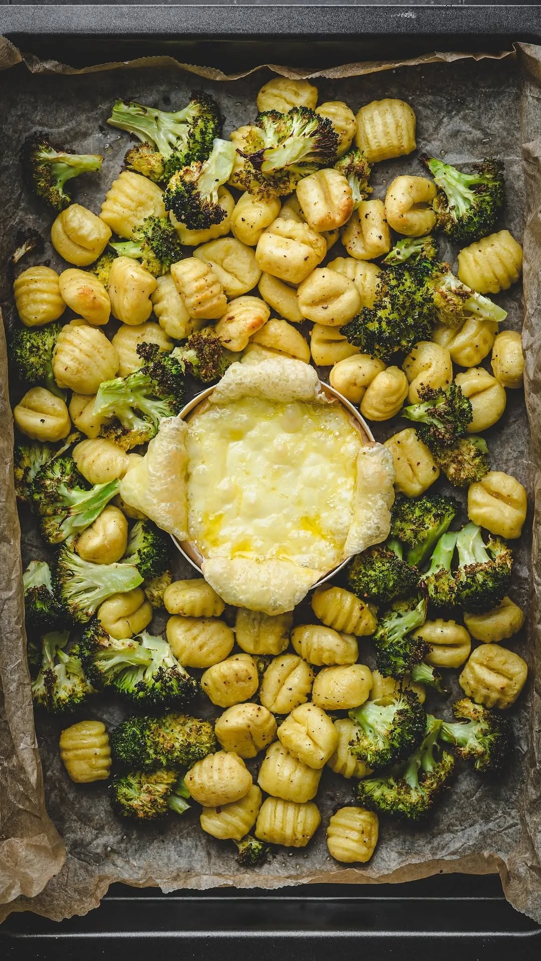 Ein mit Backpapier ausgelegtes Backblech mit gerösteten Gnocchi, Brokkoliröschen und einem mittigen Ofenkäse.