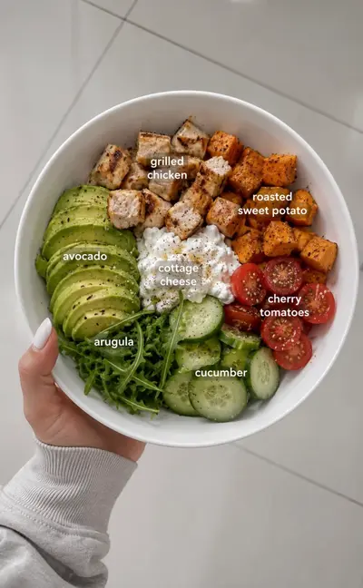Eine Hand hält eine weiße Schüssel mit einer bunten Bowl aus Hähnchen, Süßkartoffel, Hüttenkäse und frischem Gemüse.