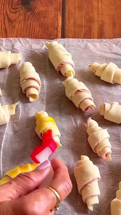 Une main badigeonne de jaune d'œuf des mini-croissants crus disposés sur une plaque recouverte de papier sulfurisé.